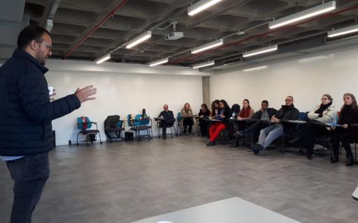 Em uma sala de aula, homem em pé fala com copo de café na mão. Ao fundo, pessoas sentadas em carteiras junto às paredes da sala ouvem e tomam notas