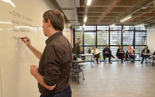 Homem escreve em quadro branco em sala de aula de universidade. Seis pessoas estão sentadas ao fundo.