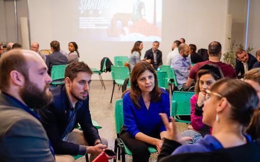 Em uma salas, grupos de pessoas conversam dispostas em círculos. Ao fundo, Powerpoint projetado em quadro branco com os dizeres "Startup POA"