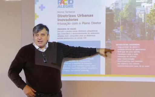 O coordenador do Pacto Alegre Luiz Carlos Pinto da Silva Filho, um homem grisalho, vestido com um blusão preto, aponta para texto em Powerpoint de apresentação