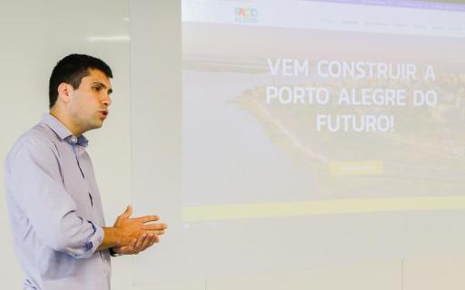 À esquerda da imagem, um homem vestindo camisa azul clara está de perfil, falando. Ao fundo, ocupando o resto da imagem, uma projeção Powerpoint mostra a capa do site do Pacto Alegre