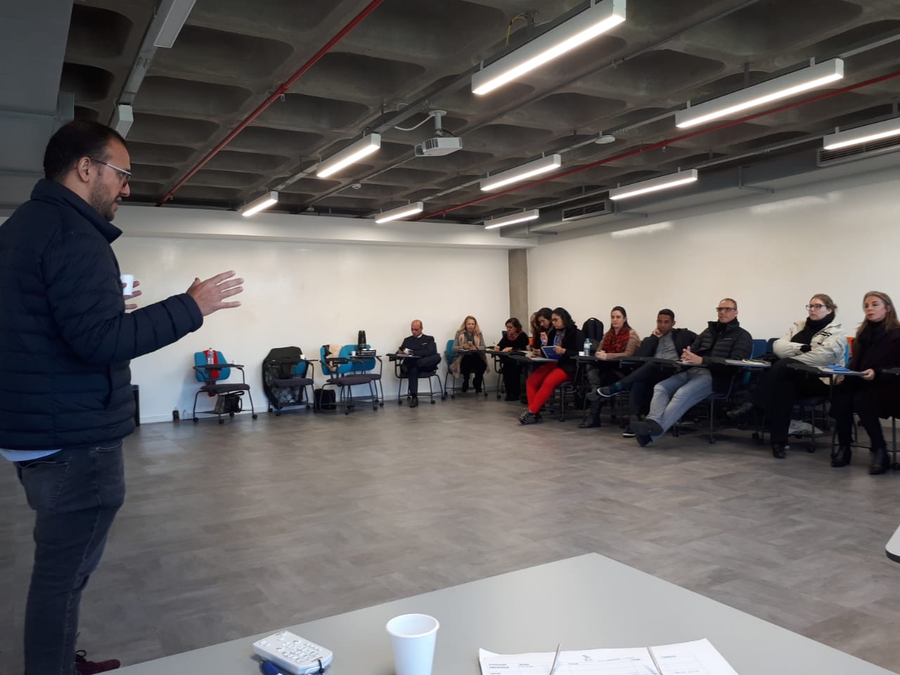 Em uma sala de aula, homem em pé fala com copo de café na mão. Ao fundo, pessoas sentadas em carteiras junto às paredes da sala ouvem e tomam notas