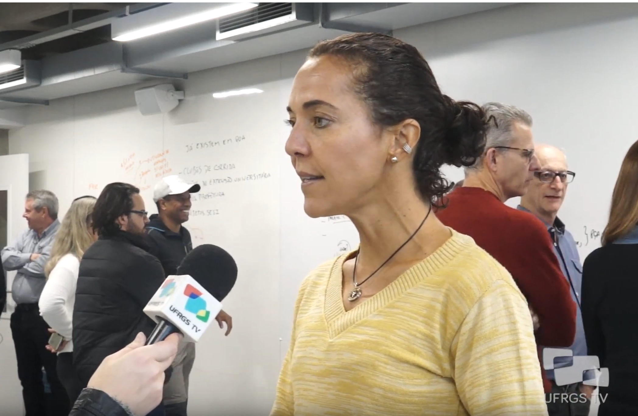 Mulher fala ao microfone de repórter. Ao fundo, grupos de pessoas conversam em frente a quadro branco