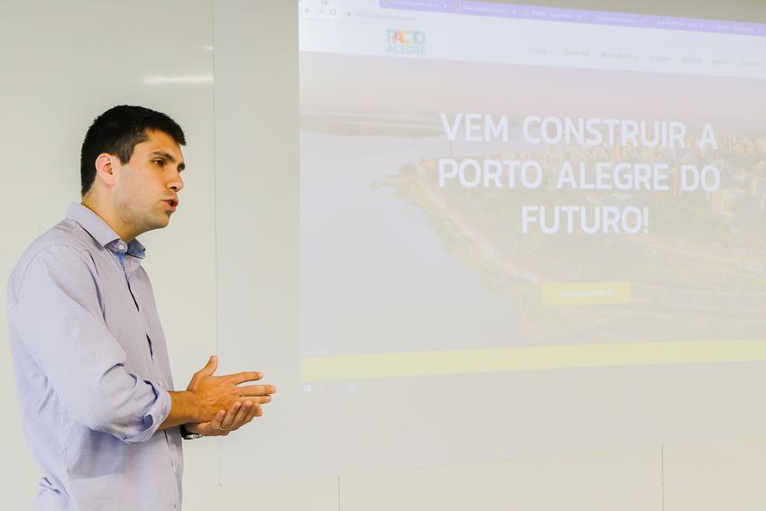 À esquerda da imagem, um homem vestindo camisa azul clara está de perfil, falando. Ao fundo, ocupando o resto da imagem, uma projeção Powerpoint mostra a capa do site do Pacto Alegre